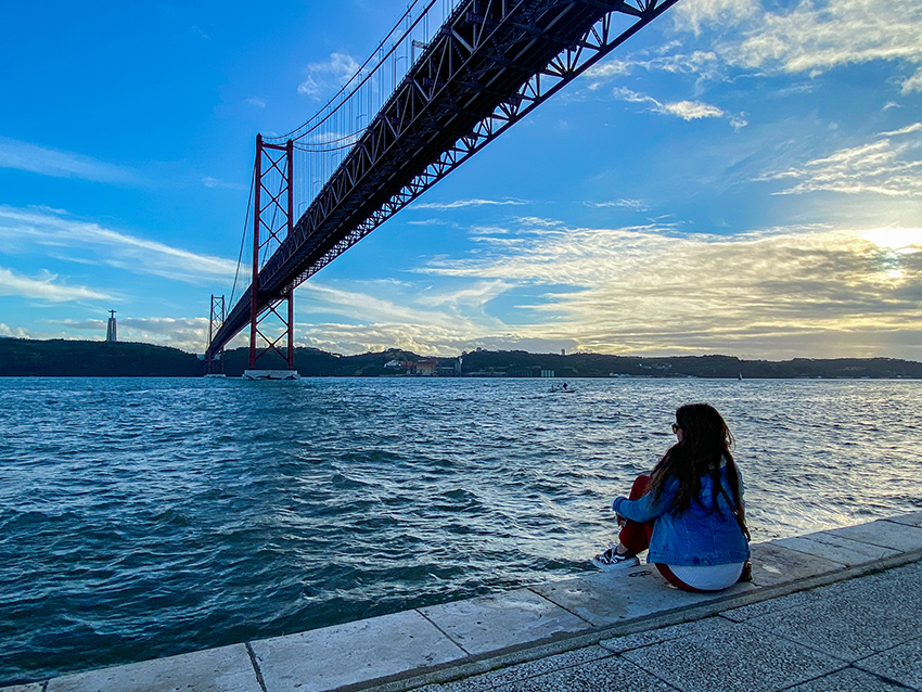 Jeune femme assise sur les bords du Tage et admirant le coucher de soleil sur le Pont du 25 avril et le Christ Roi
