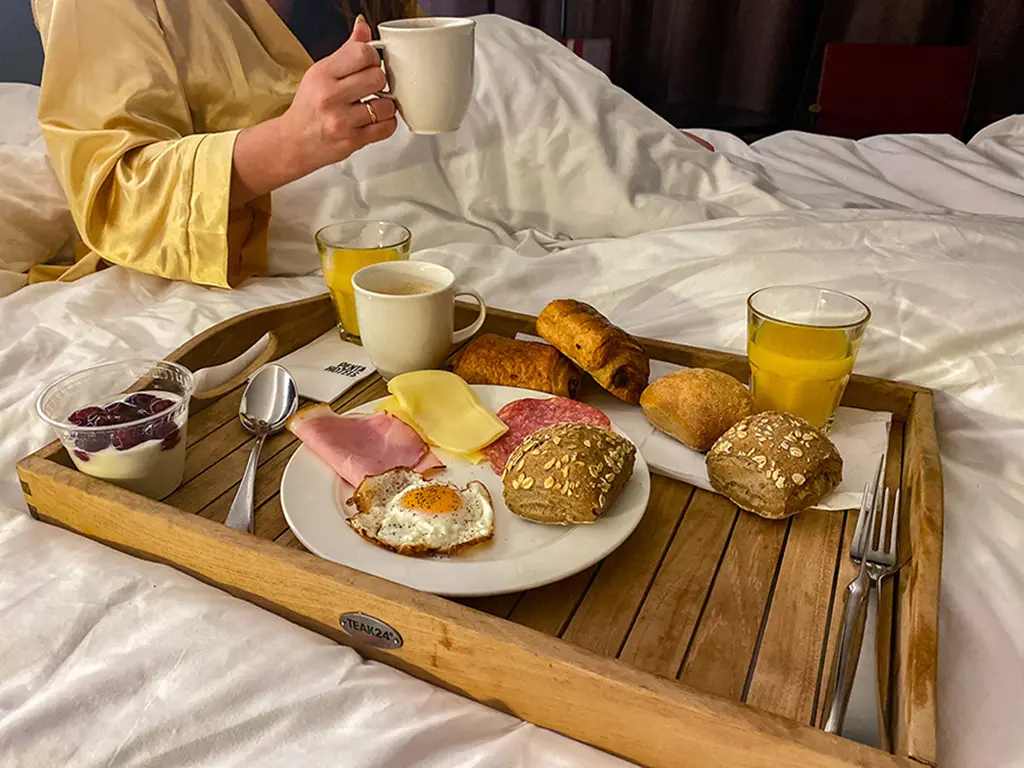 Plateau de petit-déjeuner garni de charcuterie, fromage, pain et viennoiseries, café et jus d'orange frais, ainsi qu'un yaourt aux fruits rouges. posé sur un lit