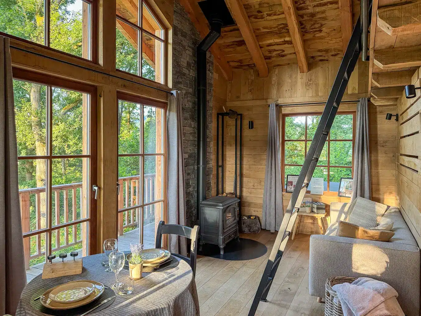 Salon du Woodpecker Lodge baigné de lumière naturelle avec grande baie vitrée, poêle à bois et coin repas, idéal pour un week-end romantique en pleine forêt.