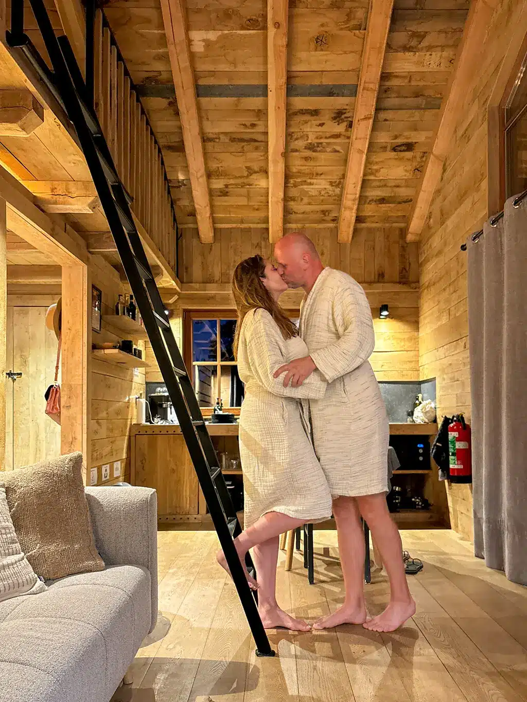 Un couple s’embrasse tendrement en peignoir au cœur d’un chalet en bois, instant complice durant un week-end romantique en Ardennes.